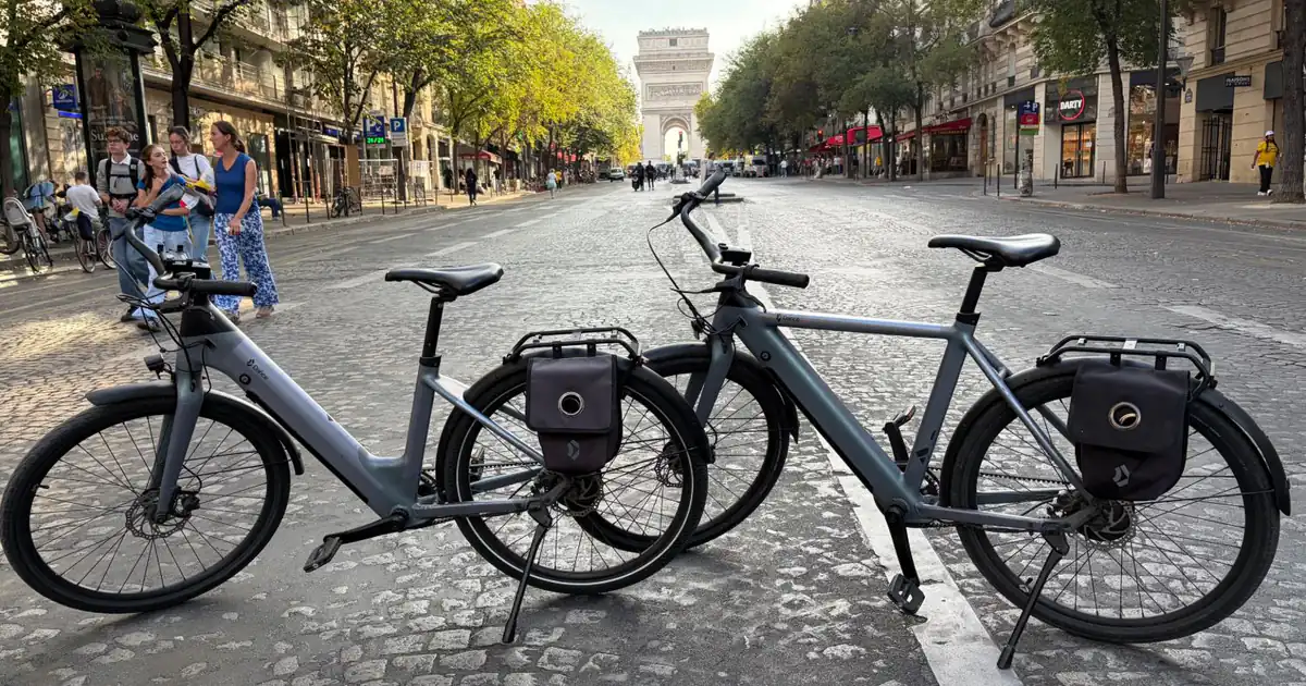 Two Dance electric bikes in the Matte Prism colour in front of the Arc de Triomphe in Paris: Dance One Step step-through on the left, Dance One high frame on the right, lock pannier bolted to each rear rack